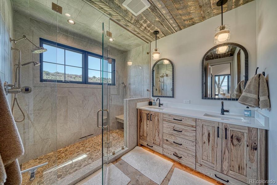 The en-suite bath features a double walk-in steam shower with frameless glass, dual vanities with rustic cabinetry, stone and tile finishes, and under-cabinet lighting. The en-suite bath features a double walk-in steam shower with frameless glass, dual vanities with rustic cabinetry, stone and tile finishes, and under-cabinet lighting.