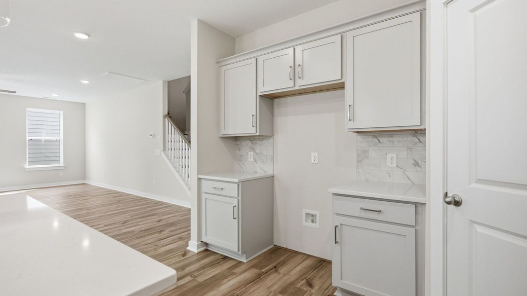 Kitchen with light cabinetry, quartz countertops, tile backsplash, and pantry with view into gathering room and staircase Kitchen with light cabinetry, quartz countertops, tile backsplash, and pantry with view into gathering room and staircase