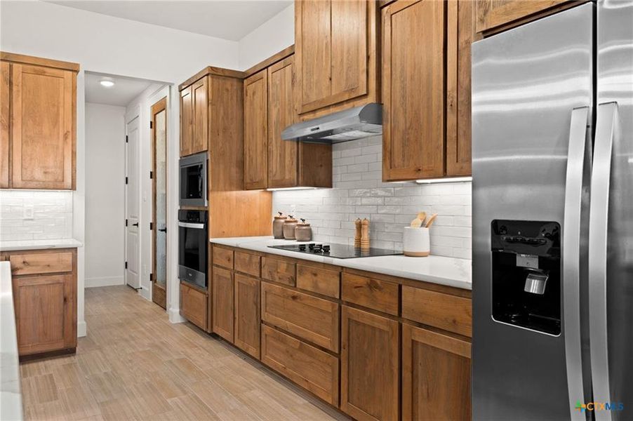 Kitchen featuring appliances with stainless steel finishes, backsplash, brown cabinetry, light wood-type flooring, and under cabinet range hood