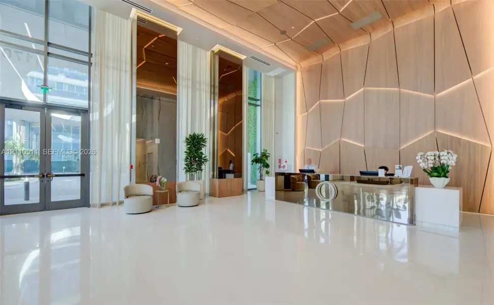 Grand lobby featuring polished white tile flooring, floor-to-ceiling glass entryway doors, geometric wood-paneled walls with integrated recessed lighting, and a mirrored reception desk