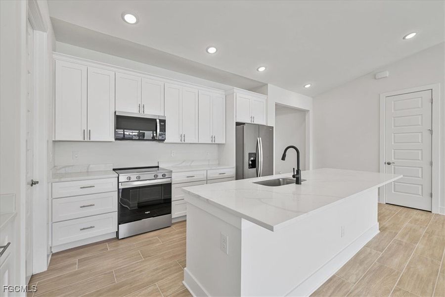 Kitchen featuring appliances with stainless steel finishes, wood tiled floors, white cabinetry, a center island with sink, and light stone countertops