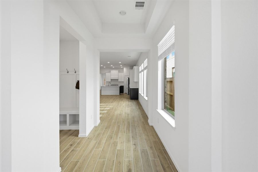 Bright, open hallway with sleek wood flooring leads to a modern kitchen, enhanced by ample natural light from large windows.