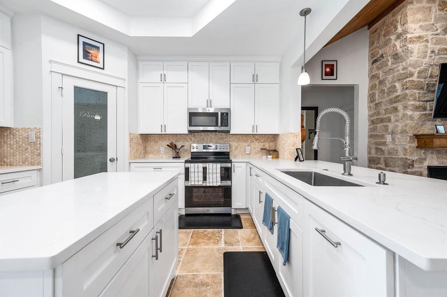 This thoughtfully designed kitchen combines modern finishes with rustic charm, featuring sleek white cabinetry, stainless steel appliances, a spacious island, and a striking stone accent wall. A large pantry provides additional storage, making the space as practical as it is stylish.