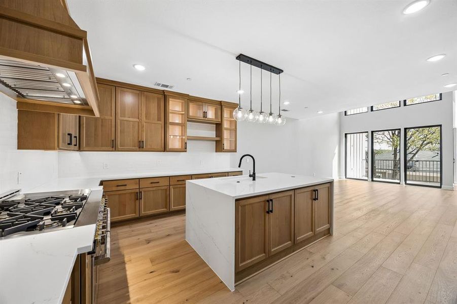 Kitchen featuring wood finish cabinets, light stone counters, an island with sink, gas stove, and open shelves Kitchen featuring wood finish cabinets, light stone counters, an island with sink, gas stove, and open shelves