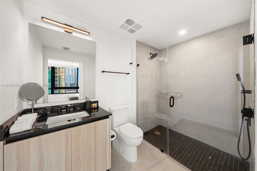 Bathroom featuring a light wood-finish vanity, dark stone-surfaced countertop, rectangular under-mount sink, matte black hardware, and a walk-in shower with a glass enclosure and dark tile flooring