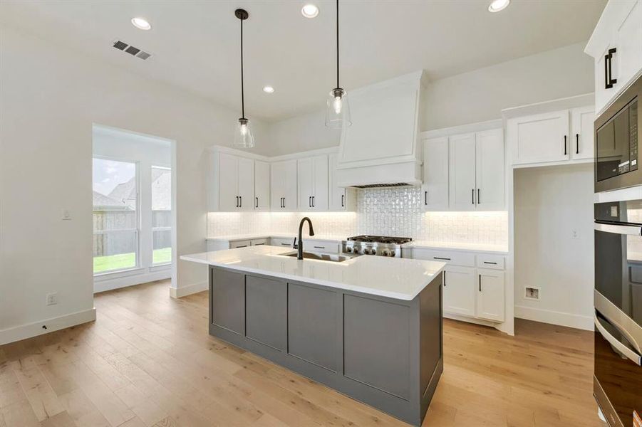 Contemporary kitchen featuring a large central island with a contrasting gray base and white countertop, white cabinetry with black hardware, a designer range hood, a tiled backsplash, and wood-finish flooring Contemporary kitchen featuring a large central island with a contrasting gray base and white countertop, white cabinetry with black hardware, a designer range hood, a tiled backsplash, and wood-finish flooring