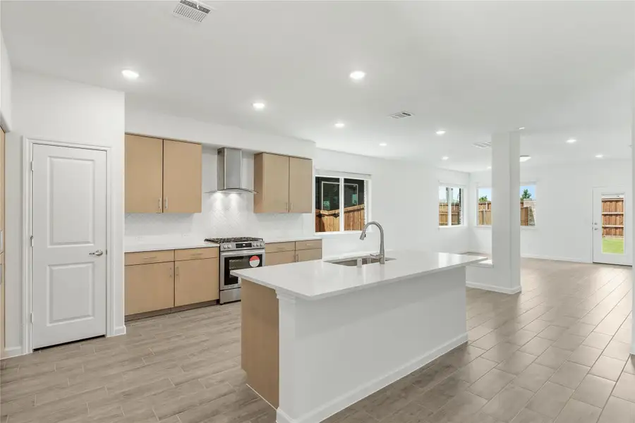 Open kitchen with white island, tan cabinets, stainless range, and wall-mount hood opening to living area