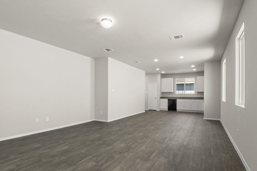 Image of home living areas with kitchen in rear with windows on one wall and over kitchen sink. There are wood-look floors and white walls