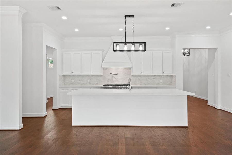 This photo showcases a modern, bright kitchen with white cabinetry, a marble backsplash, a large island with a sink, and elegant hardwood floors. The space is well-lit with recessed lighting and a chic pendant light above the island.