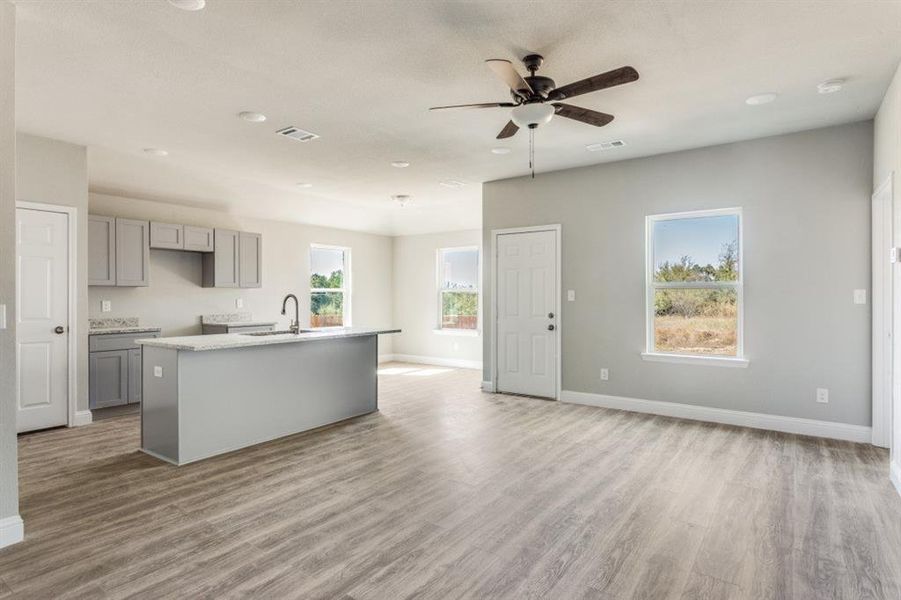 Kitchen featuring gray cabinetry, an island with sink, light wood-style floors, light stone counters, and a ceiling fan