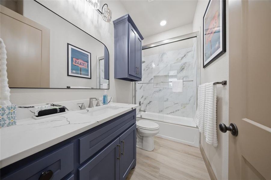 Bathroom featuring a vanity with a white countertop and blue cabinetry, a rectangular mirror, a bathtub with a clear glass sliding door, and white and gray tiled shower walls