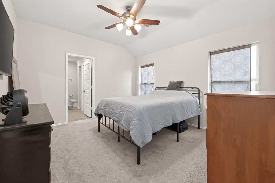 This bedroom features neutral walls and carpet, a ceiling fan, and ample natural light from two windows. It includes an ensuite bathroom for added convenience.