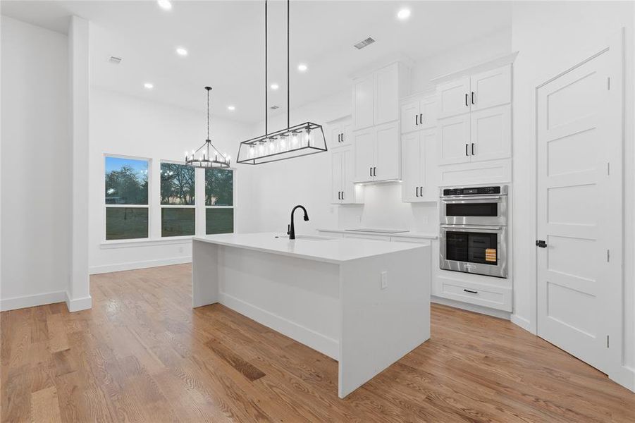 Kitchen with an island with sink, light stone counters, white cabinets, double oven, and a chandelier