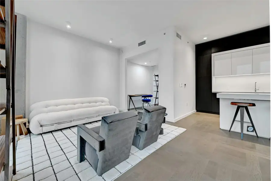This modern living area features an open floor plan that seamlessly connects to a versatile DEN, ideal for a home office, reading nook, or additional seating area. The room is bright and airy, with clean lines, neutral tones, and contemporary furnishings that create a welcoming and stylish atmosphere. The nearby kitchen is visible from the living area, offering a cohesive and functional layout perfect for both everyday living and entertaining. Furnishings provided by Scott + Cooner available for purchase separately. Photo is model unit #2406.