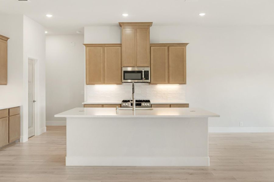 Kitchen with large white island, light wood cabinets, tile backsplash, and stainless steel appliances on light hardwood floors Kitchen with large white island, light wood cabinets, tile backsplash, and stainless steel appliances on light hardwood floors