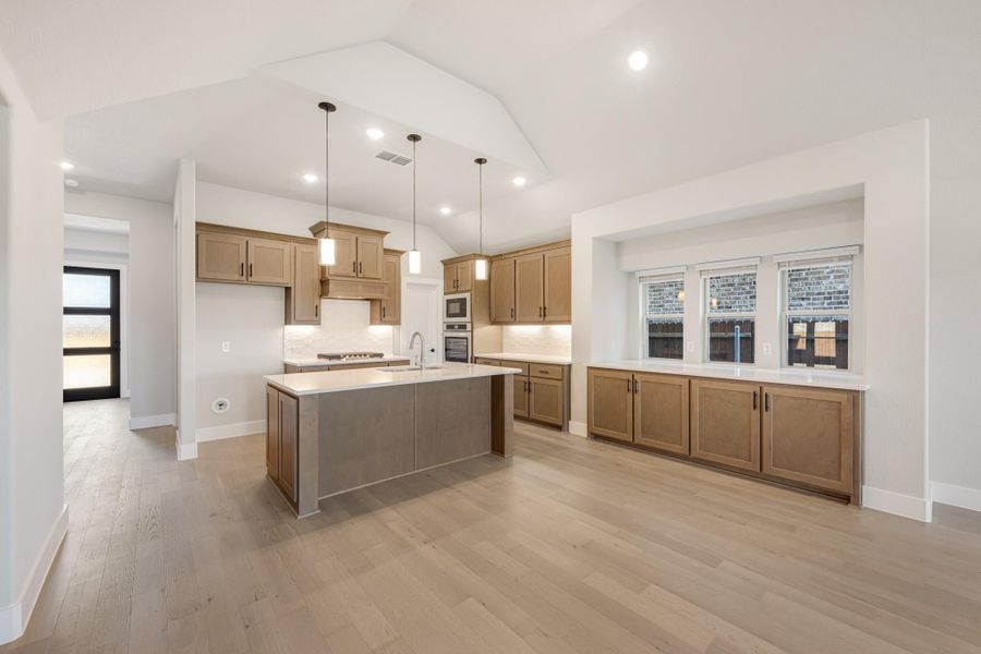 Open kitchen with wood cabinets, center island, pendant lights, and light hardwood floors under vaulted ceiling Open kitchen with wood cabinets, center island, pendant lights, and light hardwood floors under vaulted ceiling