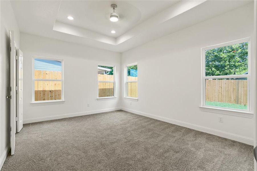 Spacious room featuring a tray ceiling with recessed lighting and a ceiling fan, plush gray carpeting, white walls, and multiple windows