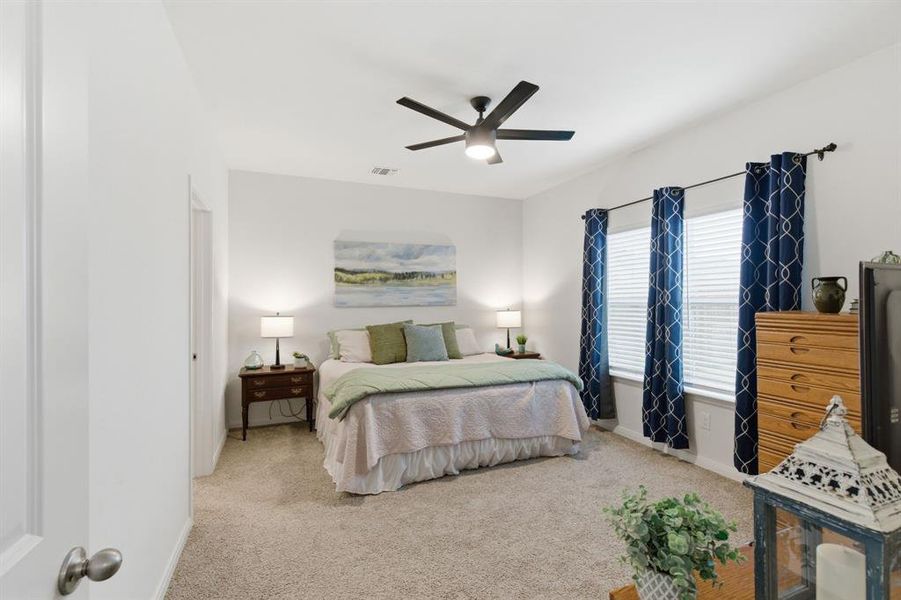 The property features a spacious room with light-colored carpet, a ceiling fan, and windows adorned with blue patterned curtains