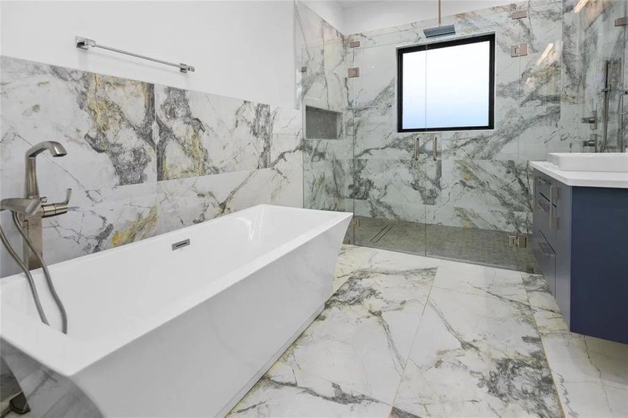 Freestanding soaking tub with a floor-mounted brushed nickel faucet, large format marble-look tile flooring and shower surround, and a glass-enclosed shower with a built-in niche Freestanding soaking tub with a floor-mounted brushed nickel faucet, large format marble-look tile flooring and shower surround, and a glass-enclosed shower with a built-in niche