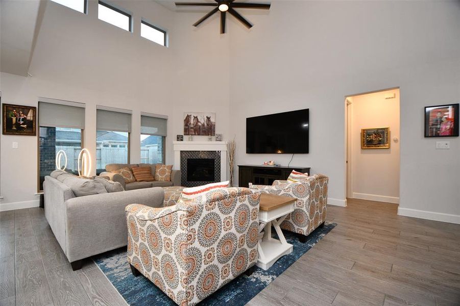 This spacious living room features high ceilings with a modern ceiling fan, abundant natural light from tall windows, and a cozy fireplace. The space is elegantly decorated with neutral tones, a large TV, and comfortable seating arrangements. Perfect for relaxation and entertainment.