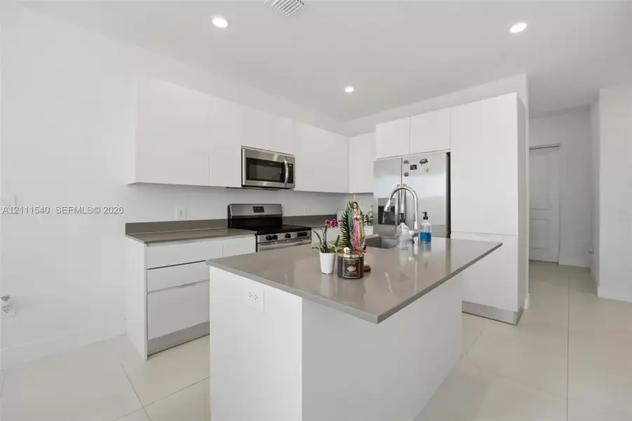 Kitchen featuring flat-panel white cabinetry, solid-surface countertops, a center island with integrated sink, stainless steel appliances, and large-format tile flooring