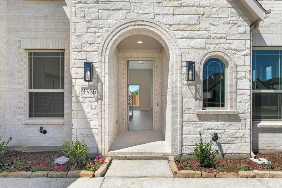 Home entrance with arched doorway, white stone exterior, wall sconces, and small flower beds at number 3316