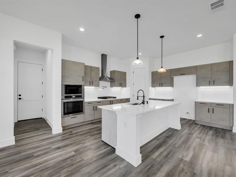 Kitchen with appliances with stainless steel finishes, wood-type flooring, an island with sink, wall chimney exhaust hood, and pendant lighting