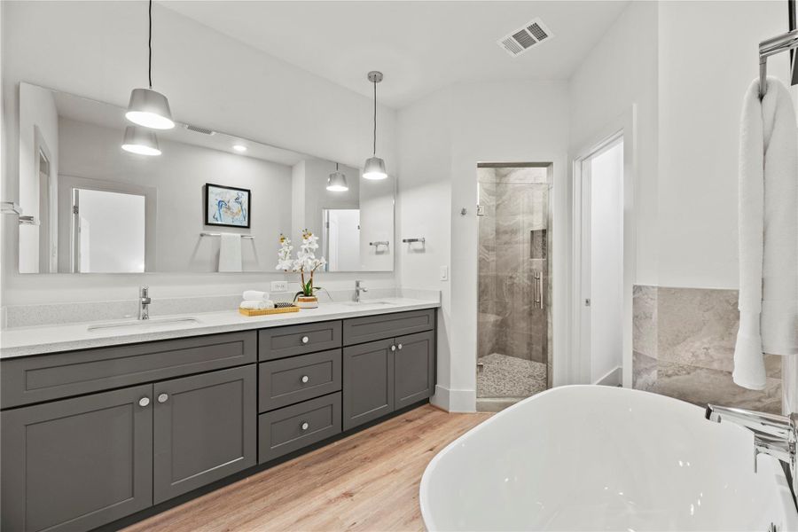 This modern primary bathroom features a spacious double vanity with sleek gray cabinets, oversized mirrors, and elegant lighting. A freestanding soaking tub and a glass-enclosed shower with stylish tile accents add a touch of luxury, all set against warm, wood-style flooring.