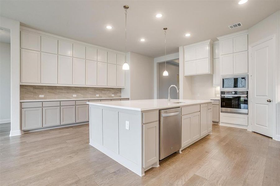 Kitchen featuring light wood-type flooring, an island with sink, decorative backsplash, stainless steel appliances, and decorative light fixtures