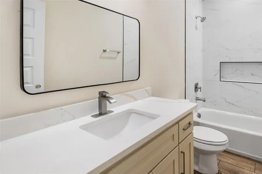 Bathroom featuring a stone countertop vanity with integrated sink, brushed nickel faucet, light wood-finish cabinetry with dark hardware, a rectangular framed mirror, and a tiled shower with a built-in niche Bathroom featuring a stone countertop vanity with integrated sink, brushed nickel faucet, light wood-finish cabinetry with dark hardware, a rectangular framed mirror, and a tiled shower with a built-in niche