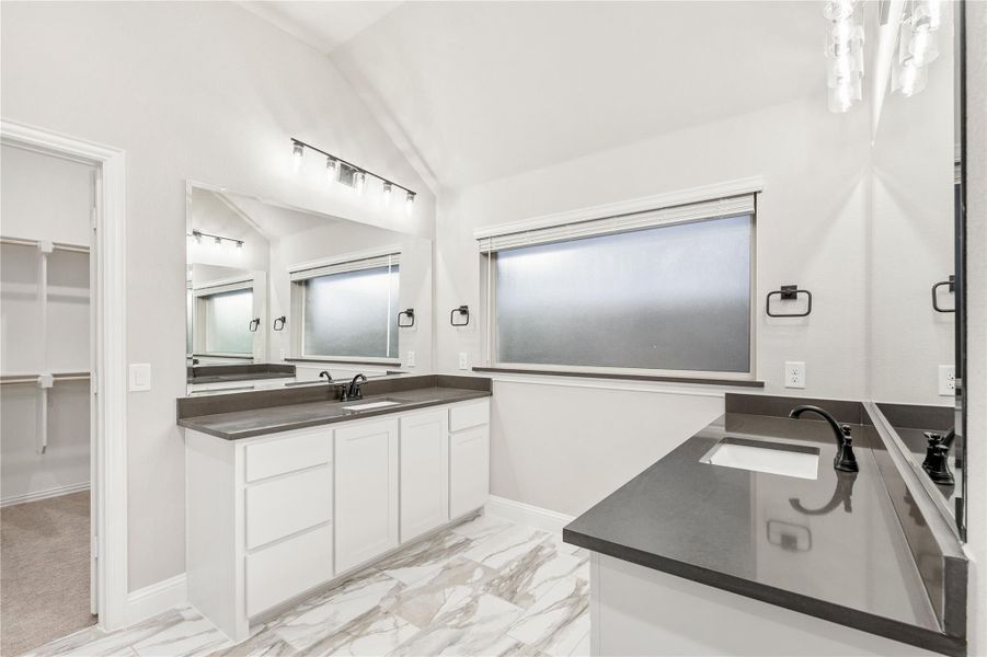 Bathroom with dual vanities, dark countertops, white cabinets, marble tile floors, and walk-in closet access