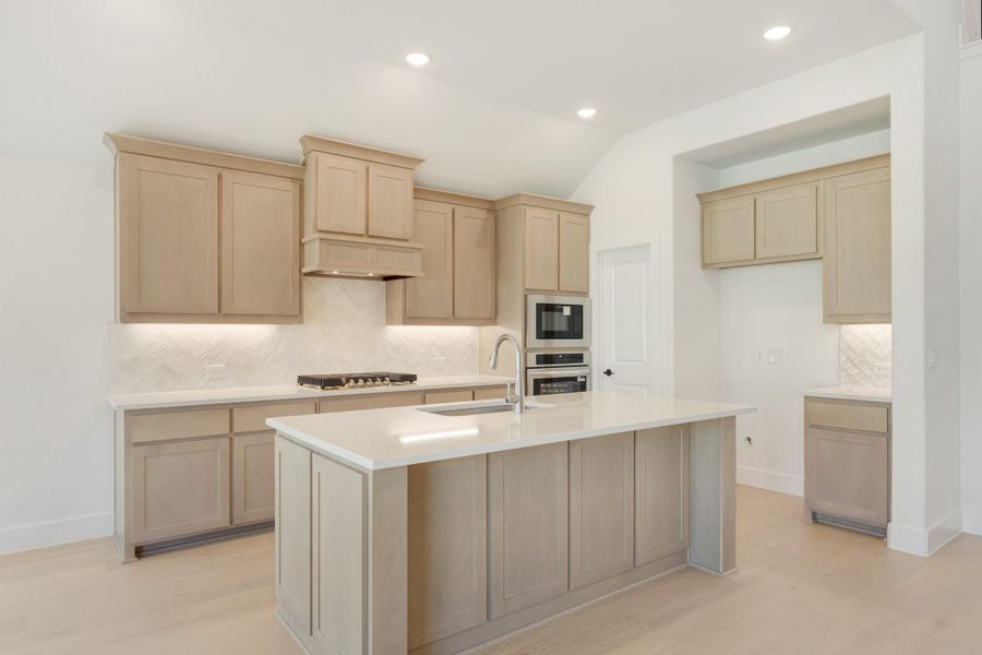Kitchen with light wood cabinets, center island with sink, stainless steel appliances, and herringbone tile backsplash