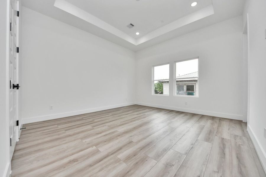 This room features a modern design with light wood flooring and a tray ceiling with recessed lighting. Two large windows provide natural light, creating a bright and airy atmosphere.