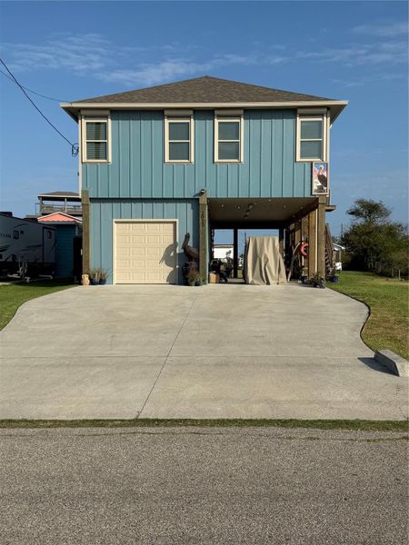 This elevated two-story home features a blue facade with a single garage and a spacious driveway. The open area underneath offers additional covered parking or storage space.