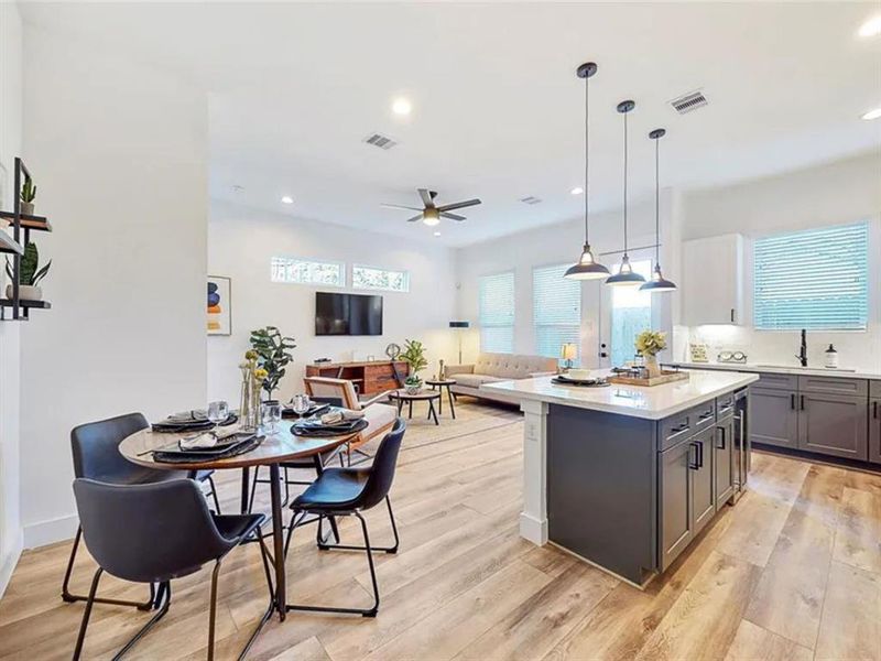 The dining area with a round wooden table and black chairs is situated in the foreground, offering a cozy space for meals. The room is bathed in natural light streaming through large windows, creating a bright and airy atmosphere. The hardwood floors throughout the space add warmth and sophistication to the overall design.