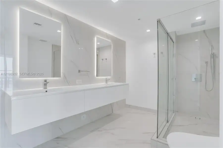 Floating dual vanity with integrated sinks, back-lit mirrors, walk-in glass shower enclosure, and full-height stone-finish wall tiling
