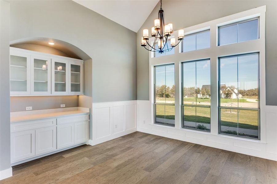 Unfurnished dining area featuring a chandelier, wainscoting, light wood-style floors, a decorative wall, and high vaulted ceiling Unfurnished dining area featuring a chandelier, wainscoting, light wood-style floors, a decorative wall, and high vaulted ceiling
