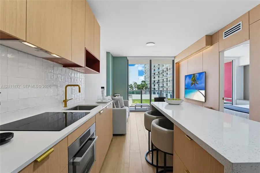 Contemporary kitchen featuring light wood-finish cabinetry, quartz countertops, a textured white tile backsplash, an integrated induction cooktop, and a matte gold faucet
