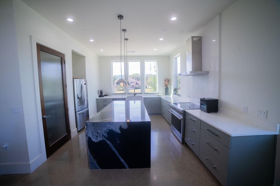 This modern kitchen features sleek gray cabinetry, a large island with a striking countertop, stainless steel appliances, and ample natural light from large windows.