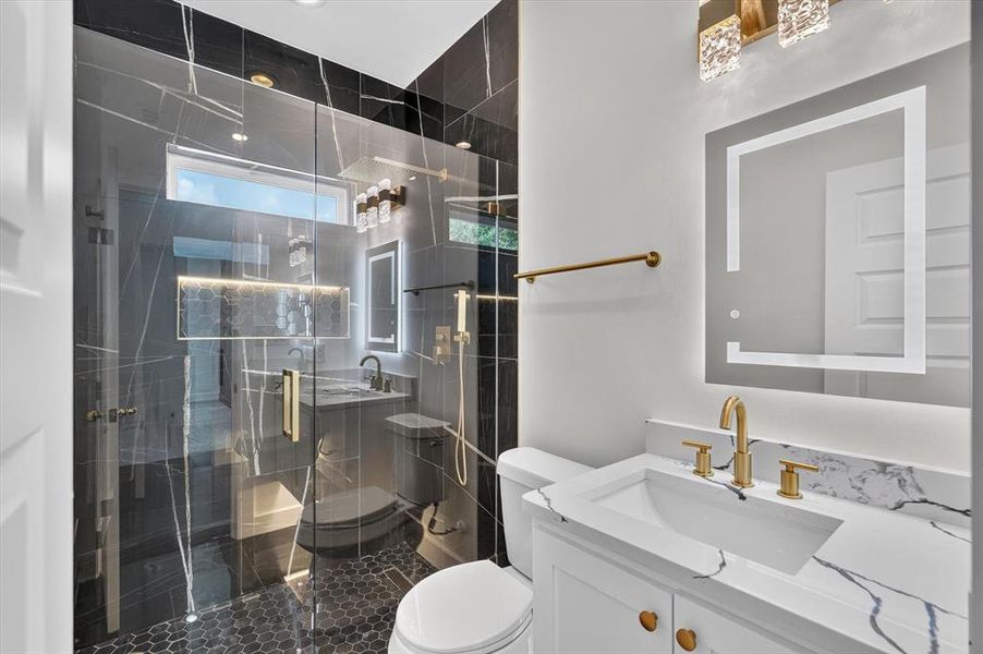 Contemporary bathroom featuring a frameless glass shower enclosure, black marble-look wall tiles, hexagonal floor tiles, a white vanity with gold-tone hardware, and an illuminated mirror
