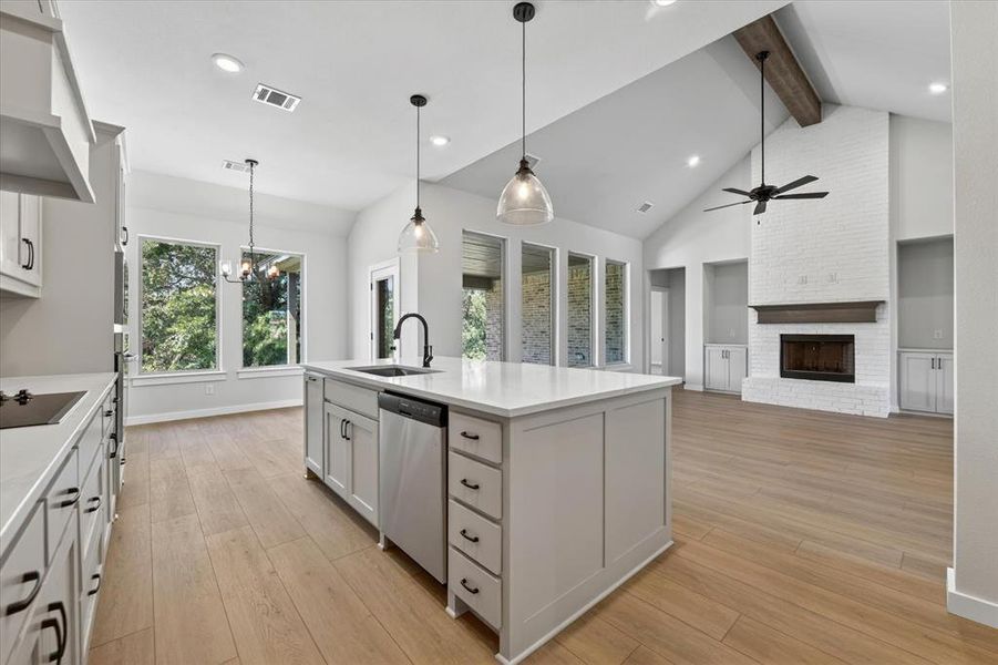 Open concept kitchen and living area featuring a central island with sink and dishwasher, wood-finish flooring, vaulted ceilings with exposed beam, and a brick fireplace with wood mantel