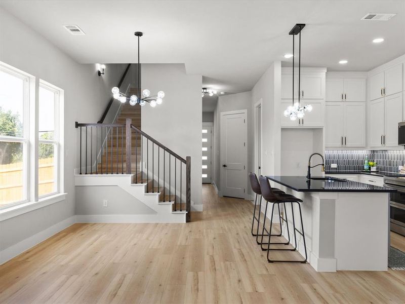 Open-concept living space featuring wood-finish flooring, a modern staircase with dark iron balusters, a bright kitchen with white cabinetry, a dark countertop island, and contemporary lighting fixtures Open-concept living space featuring wood-finish flooring, a modern staircase with dark iron balusters, a bright kitchen with white cabinetry, a dark countertop island, and contemporary lighting fixtures