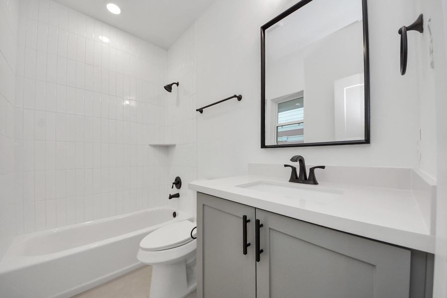 This bathroom continues the modern aesthetic with a tiled bathtub and shower combination, a black-framed mirror, and crisp, white cabinetry, providing both style and functionality.