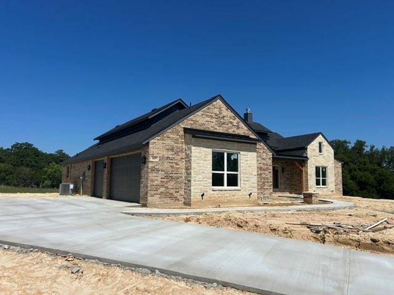 New construction featuring a multi-tone brick and stone exterior, dark shingle roofing, attached two-car garage with contemporary doors, a concrete driveway, and a covered entry with exposed wood beams