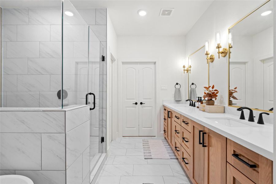 This beautifully designed primary bath showcases a large walk-in shower, quartz-topped dual vanities, custom wood cabinetry, and designer lighting that blends matte-black and gold accents for a refined, upscale feel.