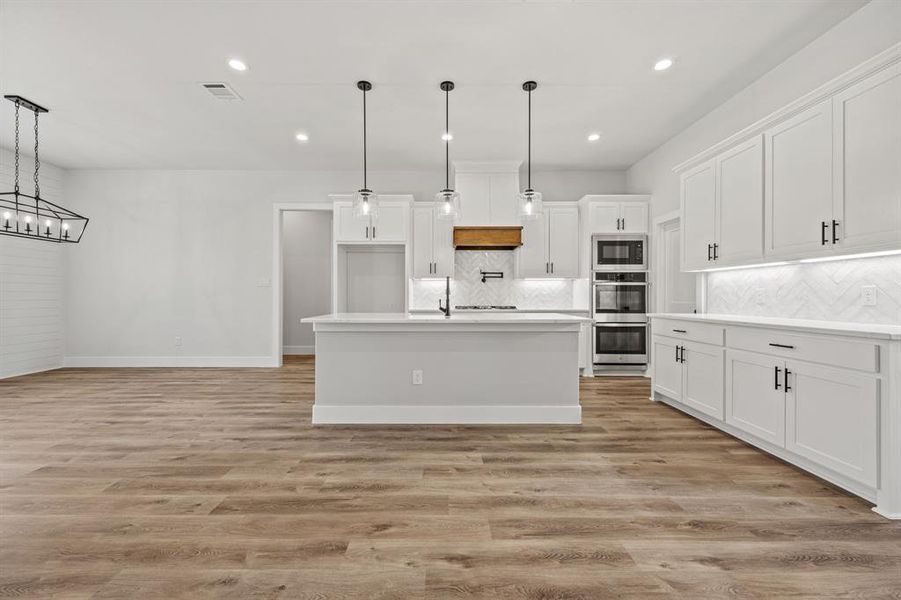 Kitchen featuring decorative backsplash, light wood-type flooring, white cabinetry, light countertops, and an island with sink Kitchen featuring decorative backsplash, light wood-type flooring, white cabinetry, light countertops, and an island with sink
