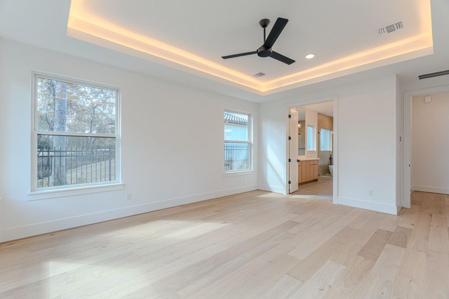 This is a spacious, bright bedroom featuring light wood floors, a ceiling fan, and recessed lighting. It offers large windows for natural light and an open doorway leading to an en-suite bathroom.