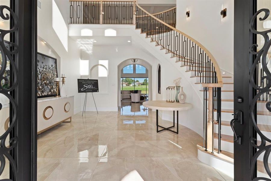 Grand foyer with high ceilings, polished floor tiles, and an elegant curved staircase with wrought-iron railings. The space is well-lit, with natural light streaming in through large windows, and features a neutral color palette that offers a sense of luxury and spaciousness.