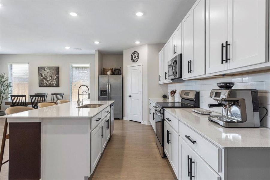 Modern kitchen featuring a large central island with an undermount sink, white cabinetry with black hardware, light-toned countertops, and light wood-finish flooring Modern kitchen featuring a large central island with an undermount sink, white cabinetry with black hardware, light-toned countertops, and light wood-finish flooring
