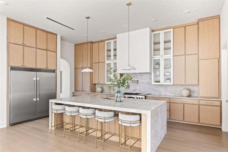 Expansive kitchen featuring a large waterfall island, light wood-finish cabinetry, stainless steel appliances, and a marble-finish backsplash